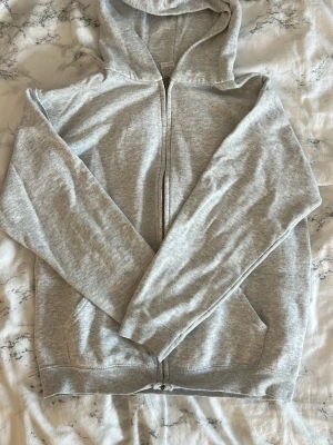 Ljusgrå hoodie med dragkedja - Klassisk ljusgrå hoodie med dragkedja framtill och två fickor. Hoodien har en skön huva och ribbade muddar vid ärmslut och nederkant. Tillverkad i mjukt bomullsmaterial för en avslappnad och bekväm stil.