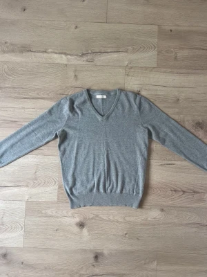 Grå stickad tröja från Berkeley - Klassisk grå stickad tröja från Berkeley i 100% bomull. Tröjan har V-ringning, långa ärmar och ribbade muddar vid ärmslut och nederkant. Perfekt för lager på lager och enkel att matcha med olika stilar.