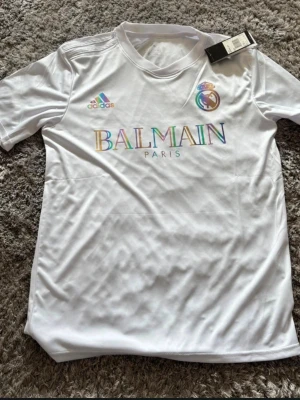 Vit Balmain x Adidas t-shirt - Säljer en vit t-shirt med Balmain Paris-tryck och Adidas-logga i regnbågsfärger. Tröjan har korta ärmar och rund hals. Materialet känns lätt och sportigt, perfekt för en clean och trendig look. Trycket är lite snett men inget man tänker på 