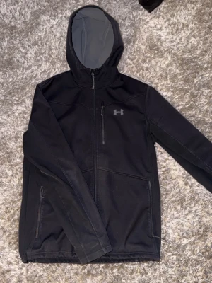 Svart vindjacka från Under Armour - Snygg svart vindjacka från Under Armour med huva och grått fleecefoder. Jackan har dragkedja framtill, tre fickor med dragkedja och diskret logga på bröstet. Perfekt för blåsiga dagar och sportiga outfits.