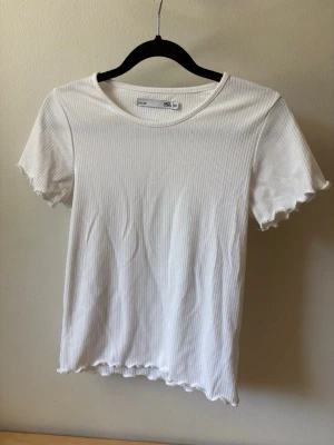 Vit ribbad topp med vågig kant - Säljer en enkel och stilren vit ribbad topp med korta ärmar och vågig kant nertill och vid ärmsluten. Toppen har en rund halsringning och är gjord i mjukt bomullsmaterial. Perfekt att matcha med jeans eller kjol.