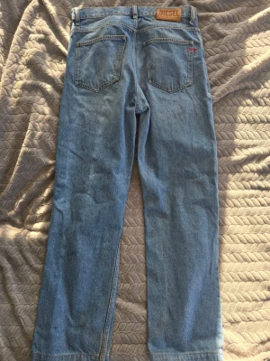 Blå raka jeans från Diesel - Säljer ett par klassiska blå jeans från Diesel med raka ben och knappgylf. Jeansen har fem fickor och en snygg vintage tvätt. Tillverkade i slitstark denim, perfekta för en avslappnad och trendig look.