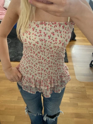 Blommig volang bandeau tubtopp - Supersöt beige-rosa tubtopp med rosa blommigt mönster. Toppen har dubbla volangdetaljer nedtill och är perfekt till sommaren. Toppen är ifrån Bikbook men jag har köpt den secondhand. Den är i storlek S men är sydd så att den är väldigt stretchig, passar nog M också.