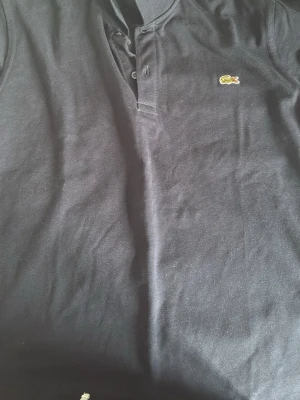 Svart pikétröja från Lacoste - Klassisk svart pikétröja från Lacoste med den ikoniska krokodilloggan broderad på bröstet. Tröjan har korta ärmar, krage och knappar framtill. Tillverkad i mjuk bomull som känns skön mot huden. Lite tight runt armarna, men sitter bra runt den