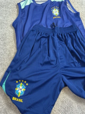 Blått Brasilien träningsset - Säljer ett blått träningsset med linne och shorts, båda med Brasilien CBF-logga och gula detaljer. Setet är gjort i ett lätt och snabbtorkande material, perfekt för fotboll eller annan sport. Elastisk midja på shortsen för skön passform.