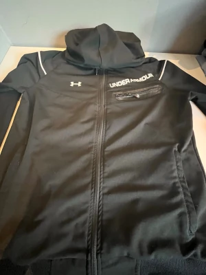 Svart hoodie från Under Armour - Säljer en svart hoodie från Under Armour med dragkedja, huva och vita detaljer på axlarna. Bröstet har en vit logga och text, samt en bröstficka med dragkedja. Materialet är en mix av bomull och polyester, vilket gör den både mjuk och sportig.