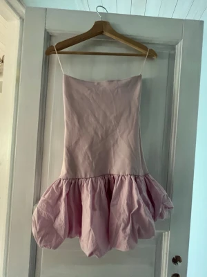 Ljusrosa kort klänning med volang - Supersöt ljusrosa kort klänning utan axelband och volang nedtill. Klänningen har en figurnära passform upptill och en fluffig kjol nertill💕