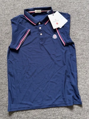 Marinblå ärmlös polotopp Moncler - Snygg marinblå ärmlös polotopp från Moncler med klassisk krage och knappar framtill. Toppens krage och ärmöppningar har rödvita ränder och ett Moncler-märke på bröstet. Perfekt för en sportig och stilren look.