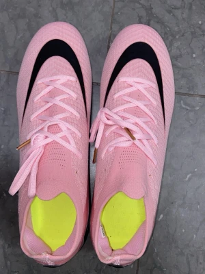 Nike Mercurial - Säljer ett par snygga fotbollsskor i rosa med svart detalj på ovansidan och gula innersulor. Skorna har snörning och plastdobbar i rosa för bra grepp på planen. Ovandelen är i ett ventilerande syntetmaterial som ger en lätt känsla och bra bollkontroll.