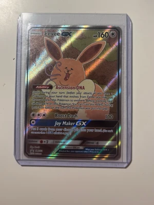 Eevee GX Pokémonkort holo - Säljer ett Eevee GX Pokémonkort med holografisk yta. Kortet har färger i brunt, rosa och silver och är inneslutet i ett genomskinligt plastfodral. Perfekt för samlare eller Pokémonfans som vill utöka sin kortsamling.