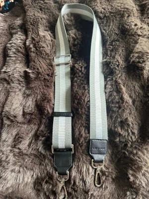 Grå axelrem från Edblad - Snygg och justerbar axelrem från Edblad i grått med ljusare ränder. Remmen är tillverkad i slitstark textil med detaljer i svart konstläder och metallspännen. Perfekt för att ge din väska en personlig touch.