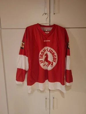 Röd IF Troja-Ljungby hockeytröja - Säljer en röd och vit hockeytröja från CCM med IF Troja-Ljungby-logga på bröstet och tryck på ärmarna. Tröjan har V-ringning och är tillverkad i ett lätt, ventilerande material som passar perfekt för ishockey eller supporterhäng.