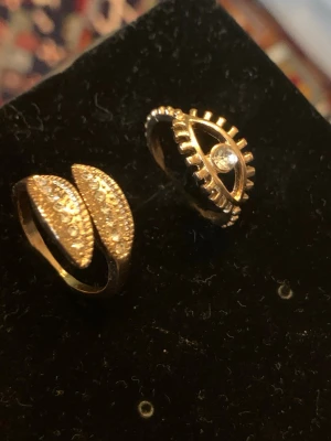Två guldiga ringar med stenar - Säljer två unika guldiga ringar. Den ena har ett ögonformat motiv med en klar sten i mitten och taggig kant. Den andra ringen har två bladformade delar täckta av små gnistrande stenar. Perfekt för dig som gillar statement-smycken och vill sticka ut.