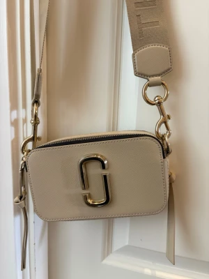 Beige axelväska Marc Jacobs - Snygg beige axelväska från Marc Jacobs med bred justerbar axelrem och guldfärgade metalldetaljer.Tillverkad i strukturerat konstläder och har en praktisk ficka på baksidan. Finns någon enstaka fläck på insidan i tyget. 