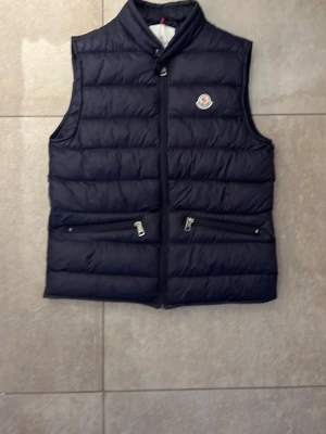 Mörkblå dunväst från Moncler - Snygg mörkblå dunväst från Moncler med hög krage och två dragkedjefickor framtill. Västen har Monclers klassiska logotyp på bröstet och quiltad design som ger en sportig och trendig look. Perfekt för lager-på-lager-stil under kyliga dagar. Du förstår på priset varför den är så billig, men genuint en otroligt snygg väst