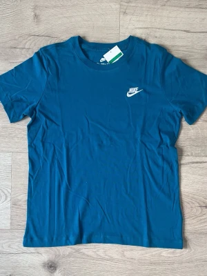 Blå Nike t-shirt i bomull - Snygg blå t-shirt från Nike med klassisk logga broderad i vitt på bröstet. Tillverkad i mjuk bomull och har en normal passform med korta ärmar och rund halsringning. Perfekt för en avslappnad och sportig stil.
