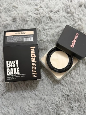 Huda Beauty Easy Bake Pound Cake puder - Löst settingpuder från Huda Beauty i nyansen Pound Cake. Kommer i en fyrkantig burk med svart lock och rosa logga. Perfekt för att fixera smink och ge en matt finish. Innehåller 20g puder i en ljus, neutral ton.