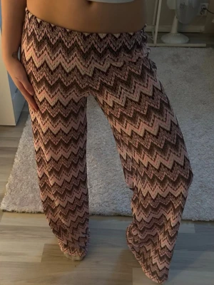 Mönstrade vida leggings i rosa och brunt - Säljer ett par snygga leggings med zigzag-mönster i rosa, brunt och beige. De har en bred resår i midjan och är vida i modellen, vilket ger en avslappnad och trendig look. Perfekta för dig som gillar färg och mönster!