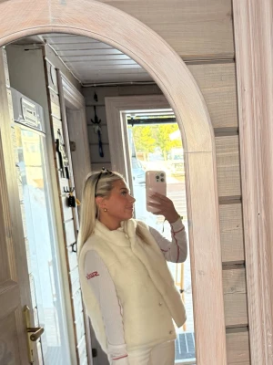 Vit Teddyväst - Snygg och fluffig vit dunväst med hög krage och tryckknappar framtill. Västen har en stilren design och är perfekt att slänga över en tröja för extra värme. Passar dig som gillar sportig och clean stil.