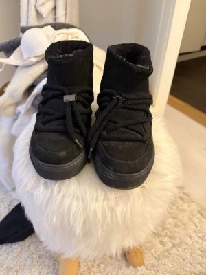 Svarta moon boots i mocka från Inuikii - Snygga svarta moon boots från Inuikii i mjuk mocka med högt skaft och breda snören som går över foten. Skorna har en chunky sula och en stilren, minimalistisk look som passar perfekt till vintern. Dragögla bak för enkel påtagning.