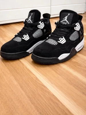 Svarta Air Jordan 4 Retro sneakers - Snygga Air Jordan 4 Retro sneakers i svart mocka med vita och svarta detaljer. Skorna har meshpaneler, vita plastdetaljer och klassisk Jumpman-logga på plösen. Tjock sula i svart och vitt ger en sportig vibe. Perfekta för dig som gillar streetwear och ikoniska sneakers.