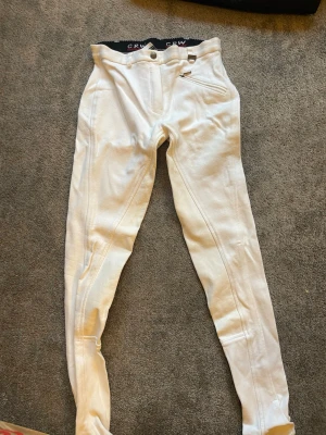 Vita ridbyxor från CRW - Säljer ett par vita ridbyxor från CRW Classic Riding Wear. Byxorna är tillverkade i 93% bomull och 7% lycra vilket gör dem stretchiga och bekväma. De har hög midja, förstärkta sömmar och en ficka fram. Perfekta för dig som gillar ridning och vill ha en klassisk look.