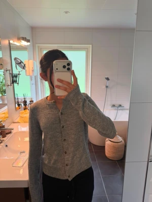 Grå kofta med knappar - Mysig grå kofta med långa ärmar och knappar framtill. Koftan har en enkel, rak passform och ribbade muddar vid ärmslut och nederkant. Perfekt att slänga på sig över en t-shirt eller linne för en chill look.