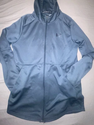 Blå zip hoodie från Under Armour - Snygg blå hoodie från Under Armour med dragkedja hela vägen och två stora fickor framtill. Tillverkad i ett mjukt och stretchigt material som känns skönt mot huden. Klassisk huva och diskret logga på bröstet. Perfekt för chill eller träning.