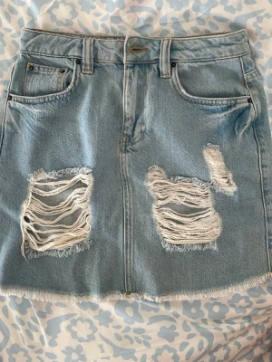Ljusblå jeanskjol med slitningar NLY - Trendig ljusblå jeanskjol från NLY Jeans med slitna detaljer framtill och rå kant nertill. Klassisk femficksmodell med bälteshällor och dragkedja. Perfekt för dig som gillar en avslappnad och cool stil.