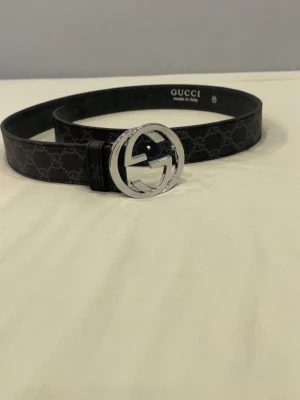 Svart  bälte med silverdetalj - Snyggt svart bälte från  med klassiskt mönster och rund silverspänne med logga. Perfekt accessoar för att lyfta din outfit. Tillverkat i skinn med exklusiv känsla och diskret mönster längs hela bältet.