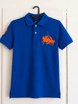 Polo Ralph Lauren blå piké med orange logga – M (10–12) 🔵🟠 - Blå pikétröja från Polo by Ralph Lauren med stor orange broderad polologga på bröstet och orange detalj på ärmen. Klassisk krage, två knappar framtill och små slitsar längst ner. Storlek: M (10–12), barn/ungdom Skick: Mycket fint skick!  Skriv gärna för mått eller fler bilder!