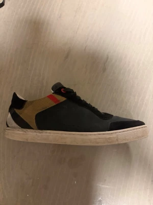 Svarta Burberry sneakers med mönster - Snygga svarta sneakers från Burberry med klassiskt rutmönster i beige, svart, vitt och rött på hälen. Skorna har svarta snören, vit sula och detaljer i mocka och textil. Perfekta för dig som vill ha en stilren men ändå unik sneaker.