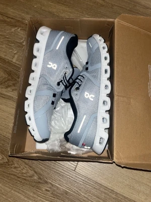 Grå On Cloud sneakers med vit sula - Snygga sneakers från On i ljusgrå mesh med vita detaljer och en unik, chunky vit sula. Skorna har svarta snören och svarta inslag vid hälen. Perfekta för dig som gillar sportig och modern stil. Reflekterande detaljer på sidan för extra synlighet.