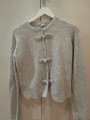 Ljusgrå kofta med rosettknäppning - Supersöt ljusgrå kofta med tre dekorativa rosetter som knäppning framtill. Perfekt för dig som gillar en gullig och stilren look.😊💕