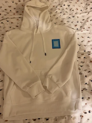 Vit hoodie  - Säljer en stilren vit hoodie med blått tryck på bröstet. Hoodien har huva med dragsko, magficka och ribbade muddar vid ärmar och nederkant. Perfekt för dig som gillar minimalistisk design och sköna material.