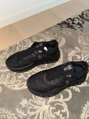 Svarta On Running sneakers - Snygga svarta sneakers från On Running med stickad ovandel och svarta snören. Skorna har en chunky sula med On:s signaturdesign och diskreta loggor på sidan och plösen. Perfekta för dig som gillar sportig och modern stil.