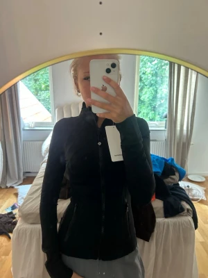 Svart träningsjacka från Lululemon - Säljer en svart, figurnära träningsjacka från Lululemon med dragkedja framtill och höga fickor. Jackan har lång ärm och hög krage, perfekt för löpning eller gym. Materialet är stretchigt och följsamt för maximal komfort.