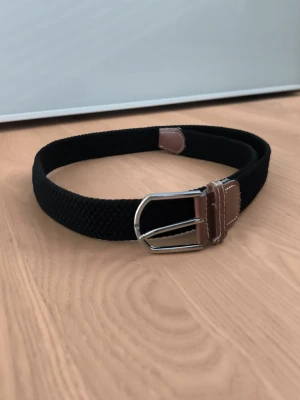Svart flätat bälte av RYVLS - Snyggt svart flätat bälte med bruna skinn-detaljer och silverfärgat spänne. 85cm långt. Från Kidsbrandstore.
