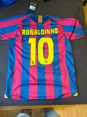 Barcelona Ronaldinho matchtröja - Säljer en klassisk FC Barcelona matchtröja från Nike med Ronaldinho och nummer 10 på ryggen. Tröjan har blå och röda vertikala ränder, gula detaljer och klubbmärke på bröstet. Perfekt för dig som älskar fotboll och vill hylla en legend. Pris kan diskuteras.