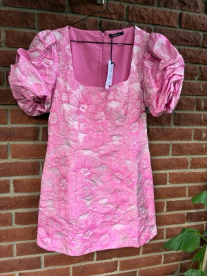Rosa blommig kortklänning med puffärm - Supersöt rosa kortklänning med stora puffärmar och fyrkantig urringning. Klänningen har ett blommigt mönster i olika rosa nyanser och är tillverkad i ett glansigt material som ger en lyxig känsla. Perfekt för alla tillfällen!
