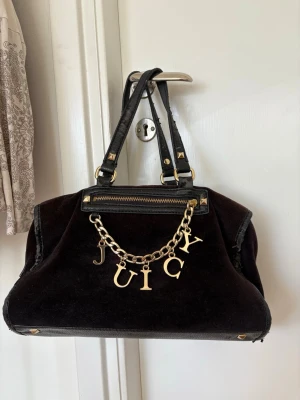 Svart handväska från Juicy Couture - Snygg svart handväska i sammet från Juicy Couture med guldfärgade metalldetaljer. Väskan har dubbla handtag och en kedja med bokstäverna JUICY framtill. 
