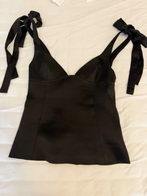 Svart satinlinnetopp från Gina Tricot - Snygg svart linnetopp i satin från Gina Tricot med smickrande passform och breda axelband som knyts till rosetter. Perfekt för att lyfta vilken outfit som helst och ger en chic vibe. Materialet har en lätt glansig finish.