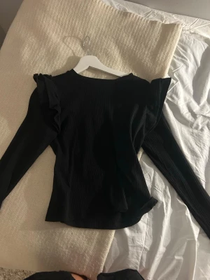 Svart ribbad topp med volang - Snygg svart ribbad topp med långa ärmar och volangdetaljer vid axlarna. Toppen har en figurnära passform och är perfekt för att lyfta din outfit med en trendig touch.