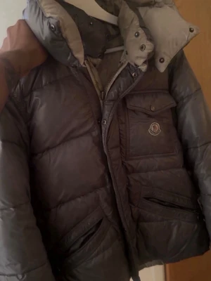 Mörkgrå pufferjacka från Moncler - Snygg mörkgrå pufferjacka från Moncler med stor huva och flera praktiska fickor framtill. Jackan har quiltad design, dragkedja och tryckknappar samt Moncler-logga på bröstet. Tillverkad i glansigt material som ger en exklusiv look.