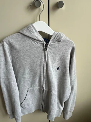 Grå hoodie från Polo Ralph Lauren - Snygg grå zip-hoodie från Polo Ralph Lauren med klassisk huva och två fickor framtill. Broderad logga i mörkblått på bröstet. Perfekt för en chill och stilren look. Mjuk bomullsblandning som är skön att ha på sig.