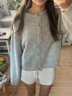 Ljusgrå mysig kofta med knappar - Ljusgrå kofta med rund hals och knäppning framtill. Koftan har en avslappnad passform, långa ärmar och ribbade muddar. Materialet ser mjukt och lite fluffigt ut, perfekt för lager på lager. Enkel och stilren design som passar till det mesta.