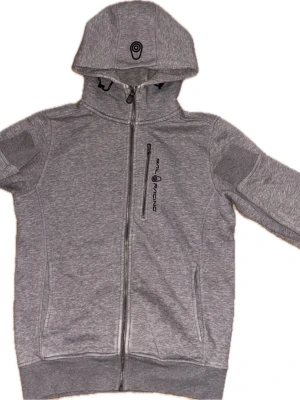 Grå sailracing hoodie med dragkedja och tryck - Snygg sailracing grå hoodie med hel dragkedja och huva. Dock dragkedjan är lite trasig men fungerar därav priset. Hör av er kring funderingar.