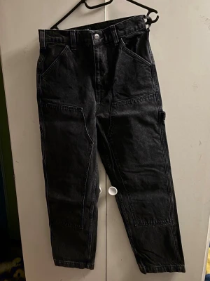 Svarta baggy jeans med utility-detaljer - Säljer ett par svarta baggy jeans med raka ben och utility-inspirerade detaljer som stora fickor fram och bak samt hammarögla på sidan. Jeansen har hög midja och breda hällor för bälte. Perfekta för en avslappnad och trendig streetwear-look.