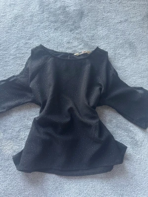 Svart topp med glittriga prickar - Snygg svart topp med glittriga små prickar och öppna axlar. Modellen har trekvartsärm och är tillverkad i ett lätt, syntetiskt material. Perfekt för dig som vill sticka ut lite extra med en trendig detalj.