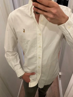 Vit skjorta från Ralph Lauren (Slim Fit) - Klassisk vit skjorta från Polo Ralph Lauren med broderad logga på bröstet. Skjortan har lång ärm, knappar framtill och button-down krage. Tillverkad i mjuk bomull för en bekväm känsla och stilren look. Storlek S slim fit. Snabb leverans!